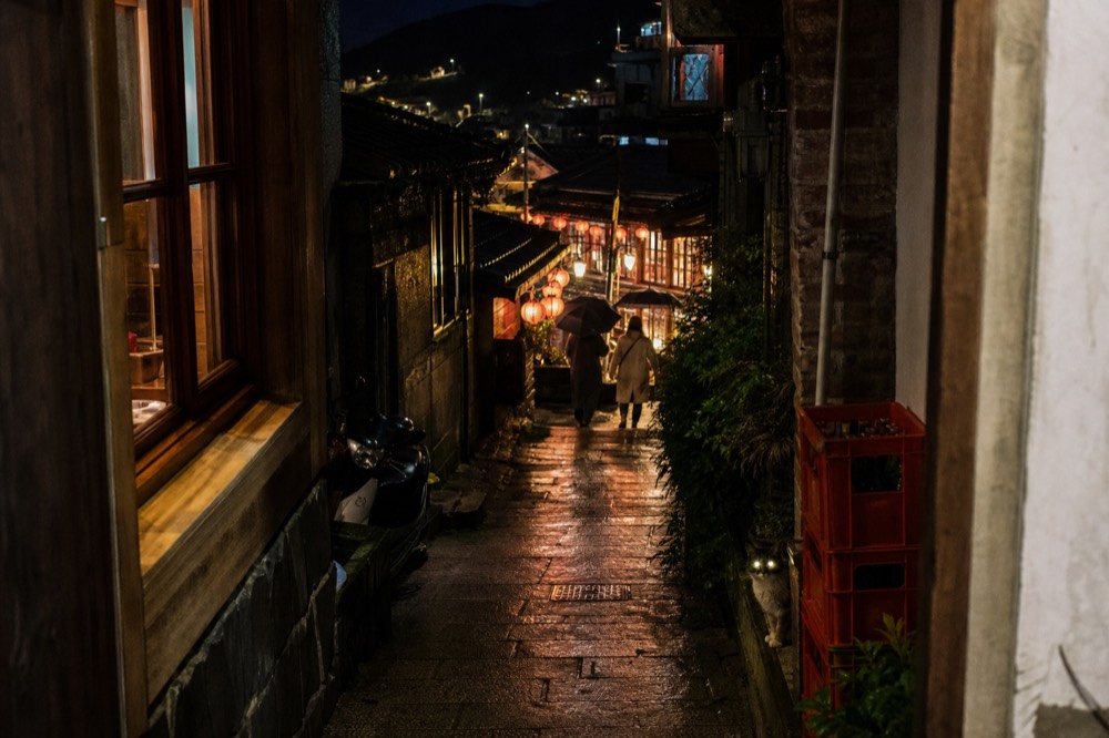 Narrow alley between old buildings with warm lamplight glowing in doorways and windows