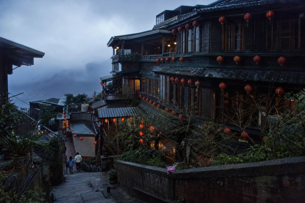 Traditional teahouses with dark wooden facades and red lanterns stacked up a steep hillside above the sea at dusk
