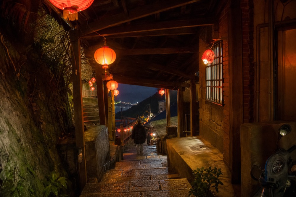 Red lanterns glowing along a covered stairway lane between old buildings after dark