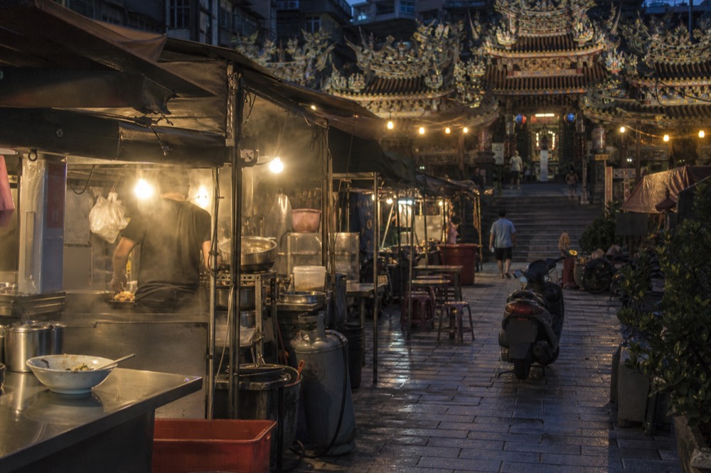 Night market stalls glowing under lanterns beside a temple in a harbour city