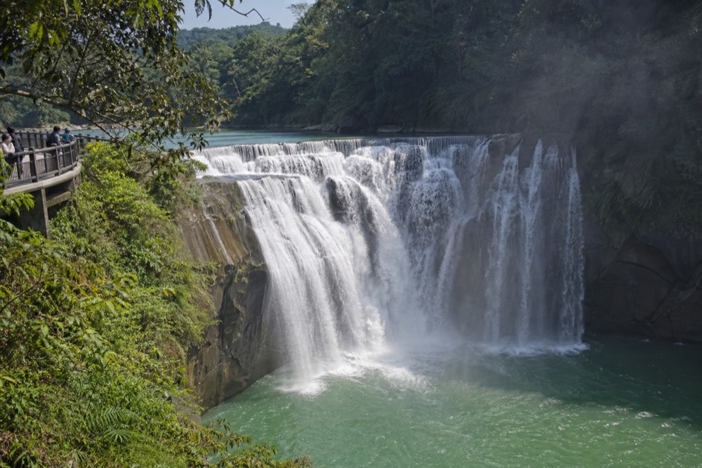 A wide waterfall dropping in a curtain across a forested river gorge