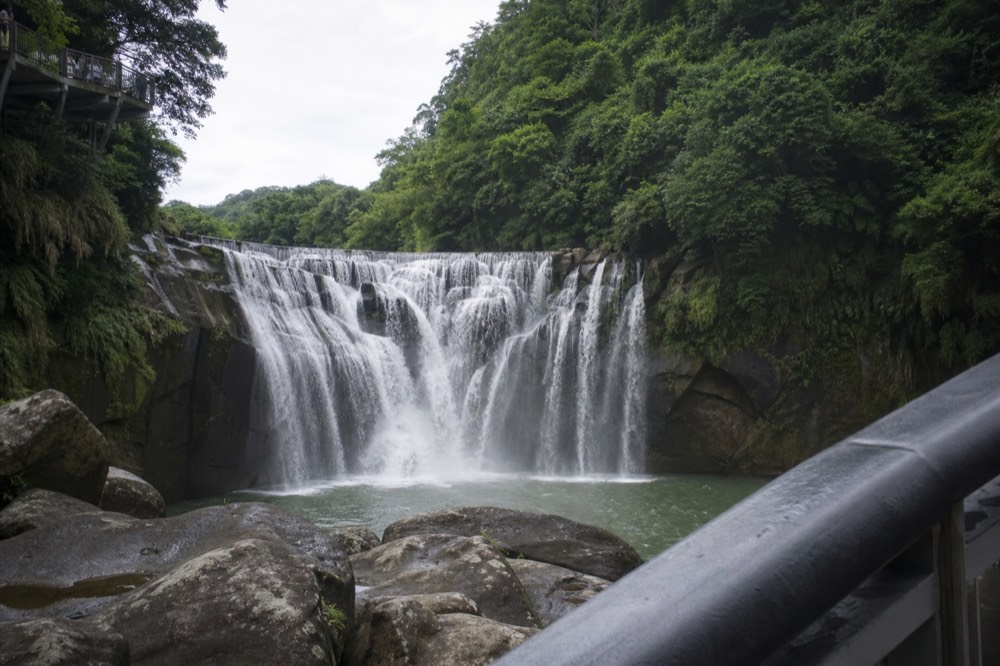 A wide waterfall spreading across a rock ledge into a misty pool, framed by green forested slopes