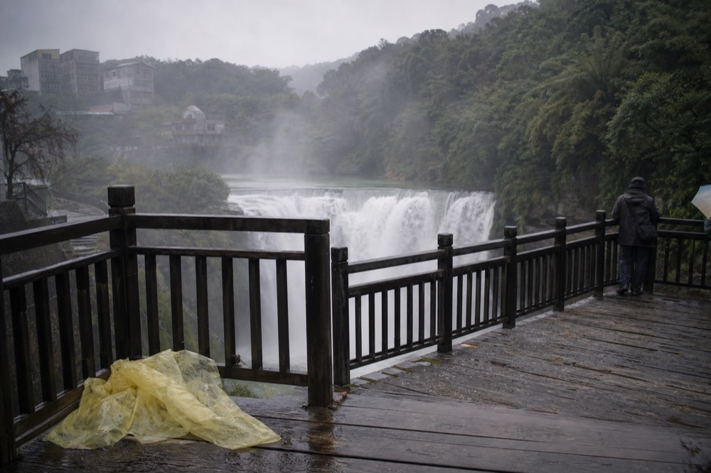 A waterfall pouring in a wide sheet over a rock lip, seen from a wooden riverside viewing platform