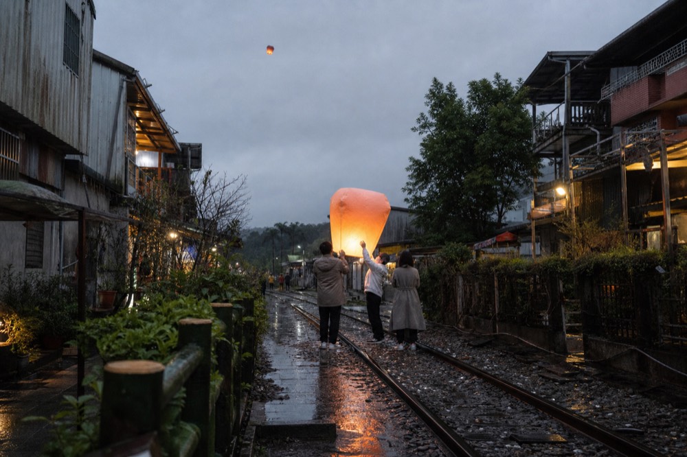 Groups of visitors standing on railway tracks beside old street shopfronts releasing a glowing paper lantern into the sky