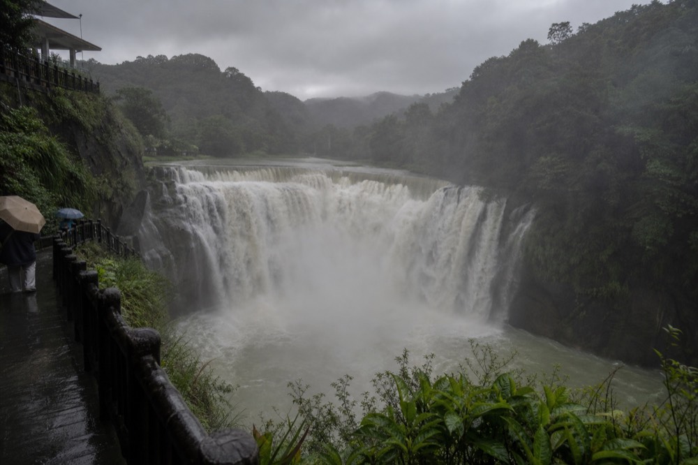 A wide waterfall in full flow filling a gorge with mist, with dark rain clouds over forested ridges