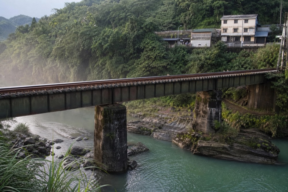 A railway bridge crossing a green river in a steep valley, with old street buildings along the far bank