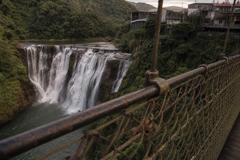 A wide waterfall seen from a suspension bridge, white water spreading across a dark rock face in a green gorge