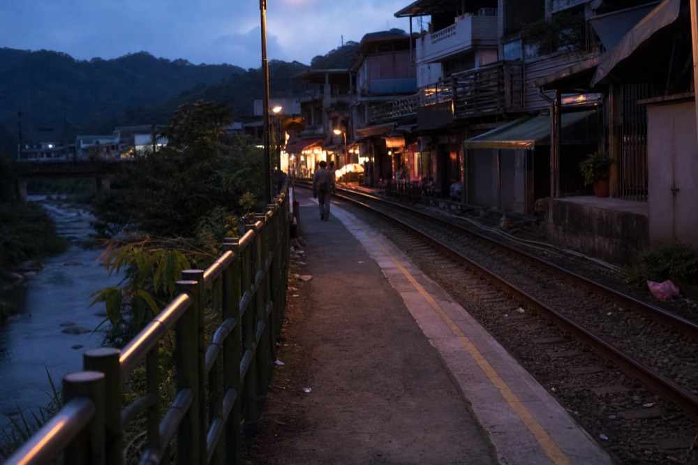 A quiet railway platform beside a river at dusk, with lantern-lit shopfronts glowing along the old street beyond
