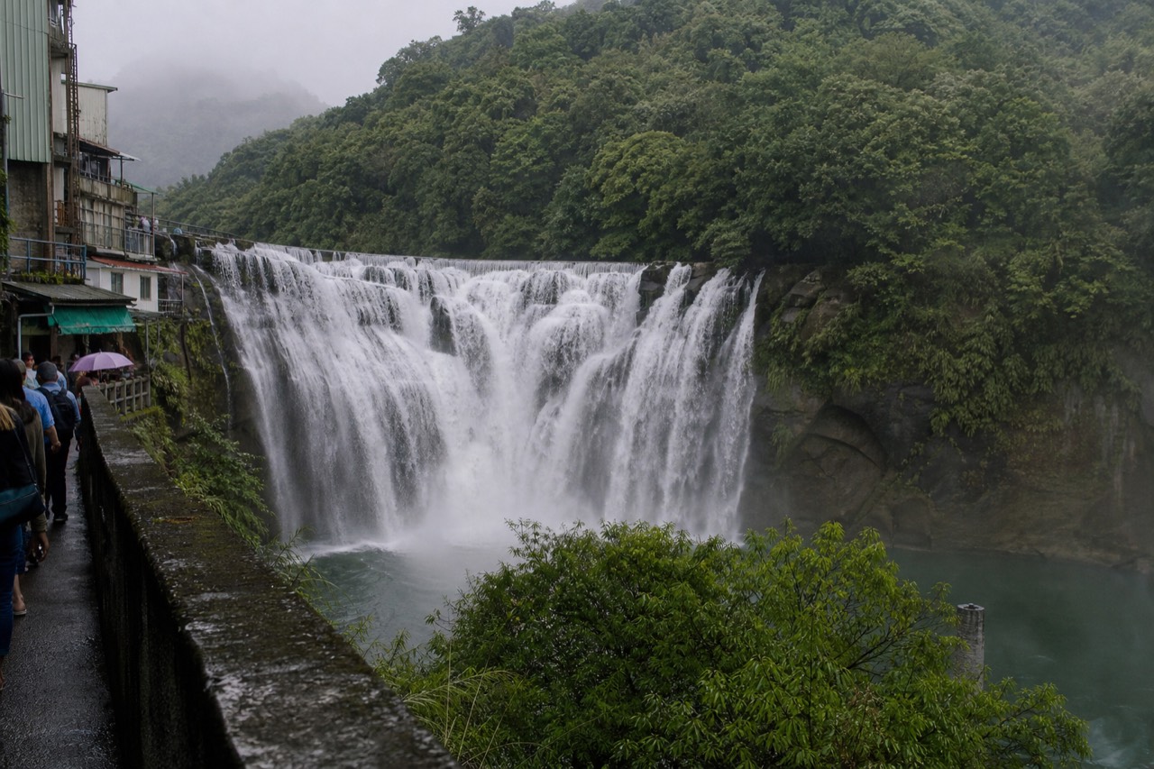 A wide waterfall dropping in a white curtain across a rock face above a forested valley in northeast Taiwan
