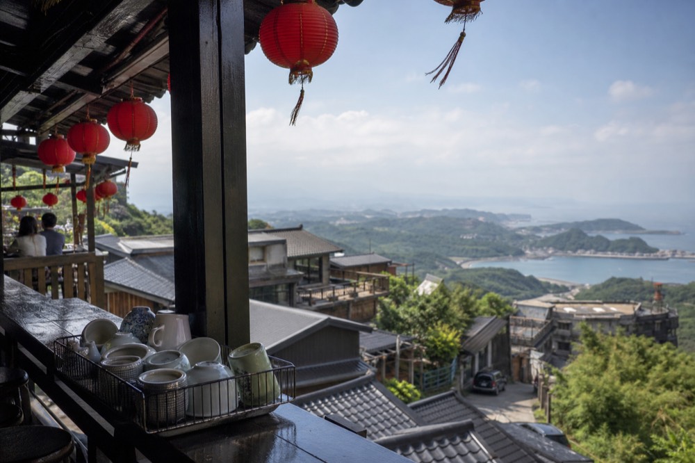 A wooden teahouse balcony overlooking a coastal mountain town and the sea beyond
