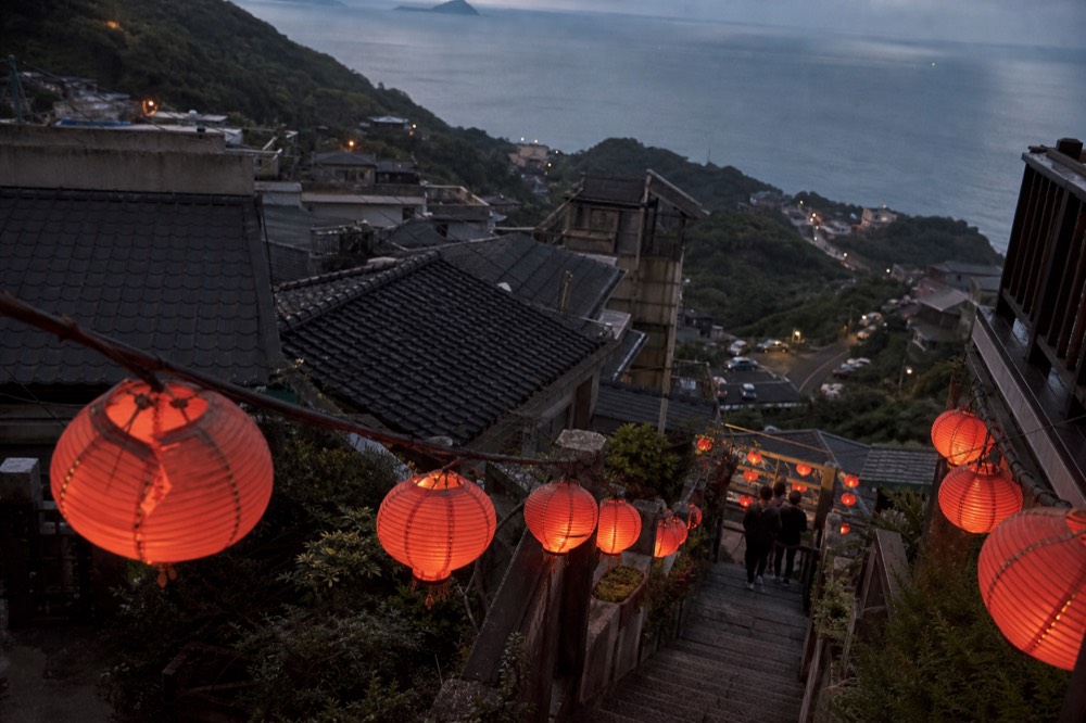 A hillside town at dusk with red lanterns glowing along its lanes and darkening sea beyond