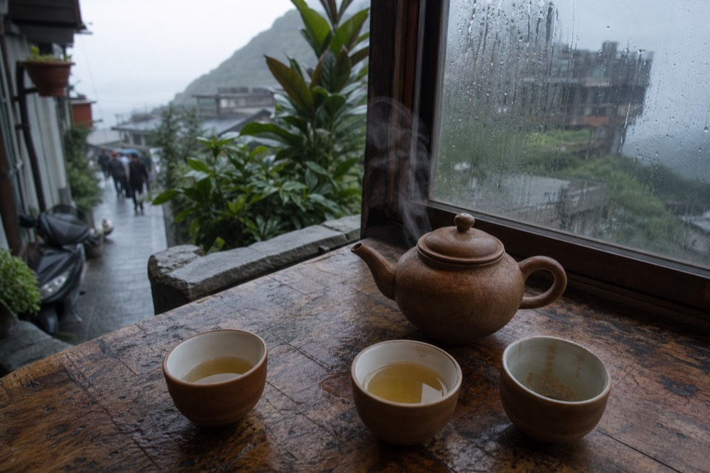 A steaming clay teapot and small cups arranged on a worn wooden table beside a window