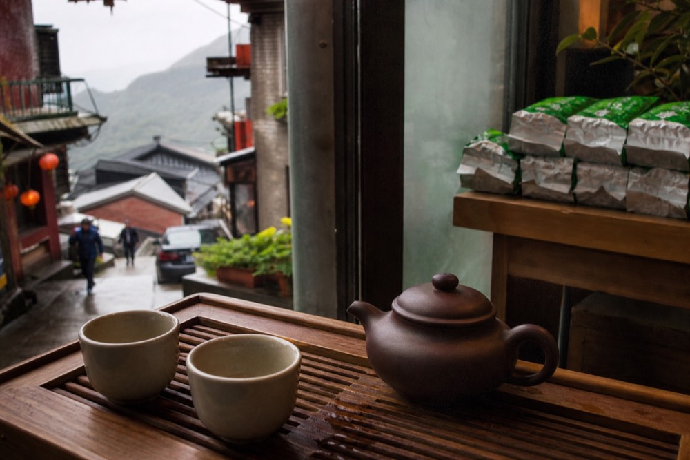 A tea set with a clay pot and small cups on a wooden tray, with Jiufen's hillside in the background
