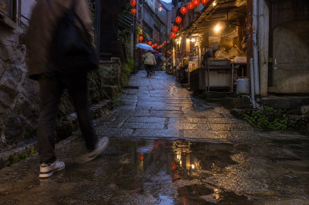 A narrow alley in Jiufen with red lanterns and food stall signs, wet cobblestones after rain
