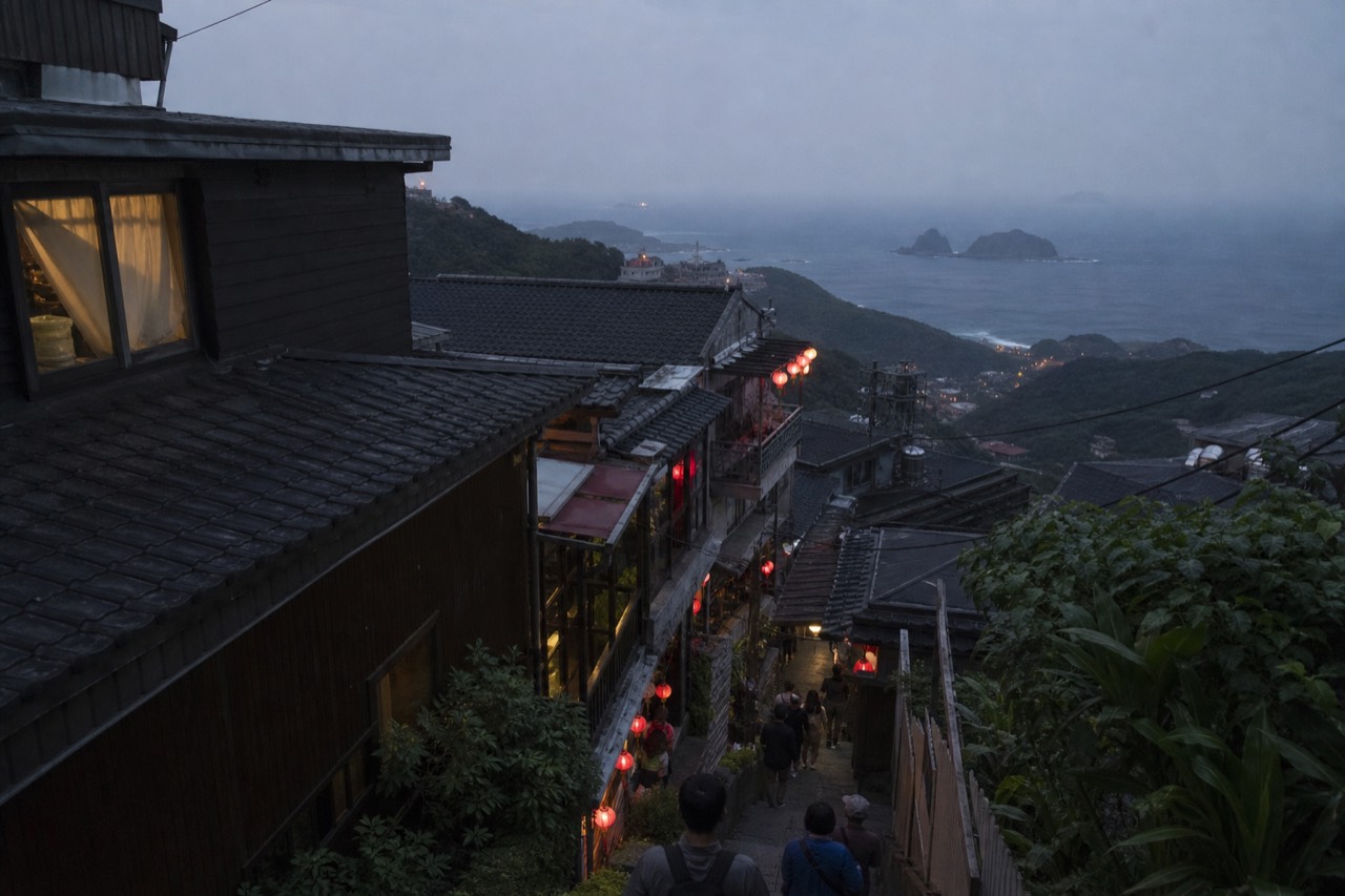 Red lanterns and tea house rooftops on a hillside street above the sea at dusk
