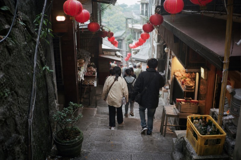 A steep Jiufen lane with red lanterns overhead and shop stalls on both sides, visitors walking the stone steps